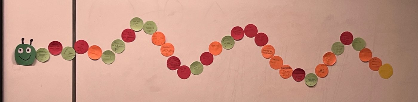 A long, wavy caterpillar made of green, red, orange, and yellow paper circles taped across a whiteboard. Each circle has handwritten encouragement on it. The caterpillar has a smiling green paper head with two antennae.